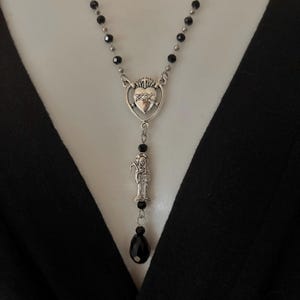 Puede incluir: Collar plateado con un colgante de coraz&oacute;n, una figura de santo y cuentas negras. El collar tiene un dise&ntilde;o de colgante de gota y una cuenta negra en forma de l&aacute;grima.