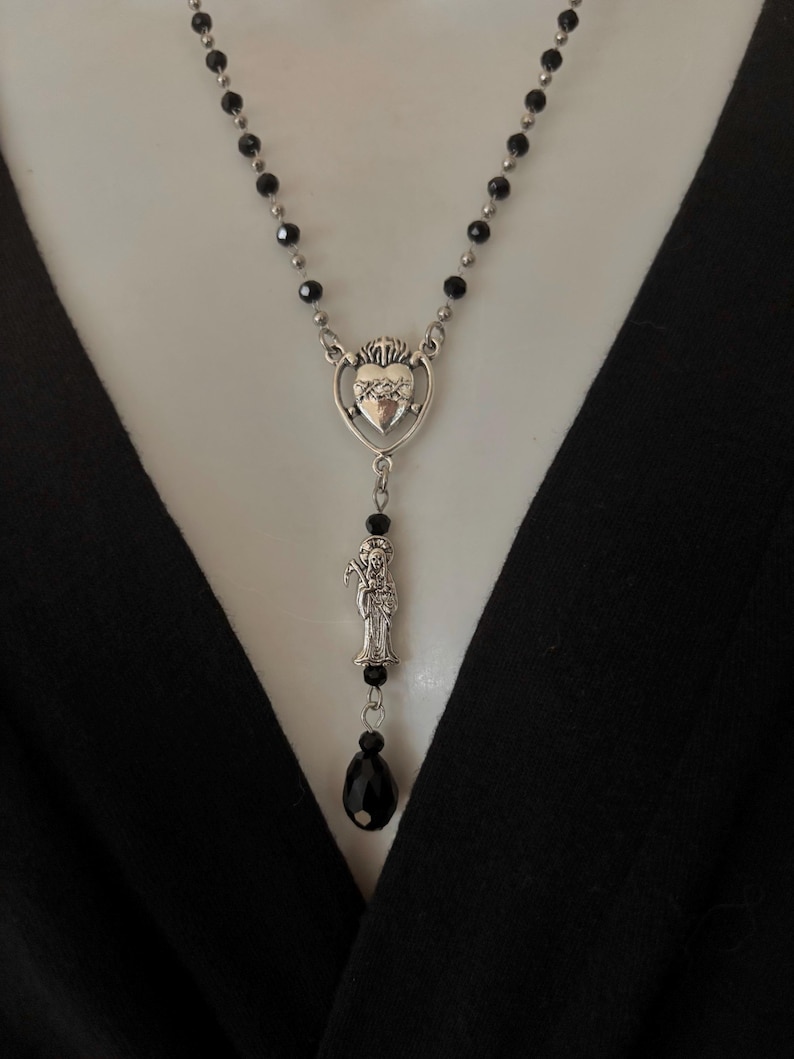 Puede incluir: Collar plateado con un colgante de coraz&oacute;n, una figura de santo y cuentas negras. El collar tiene un dise&ntilde;o de colgante de gota y una cuenta negra en forma de l&aacute;grima.