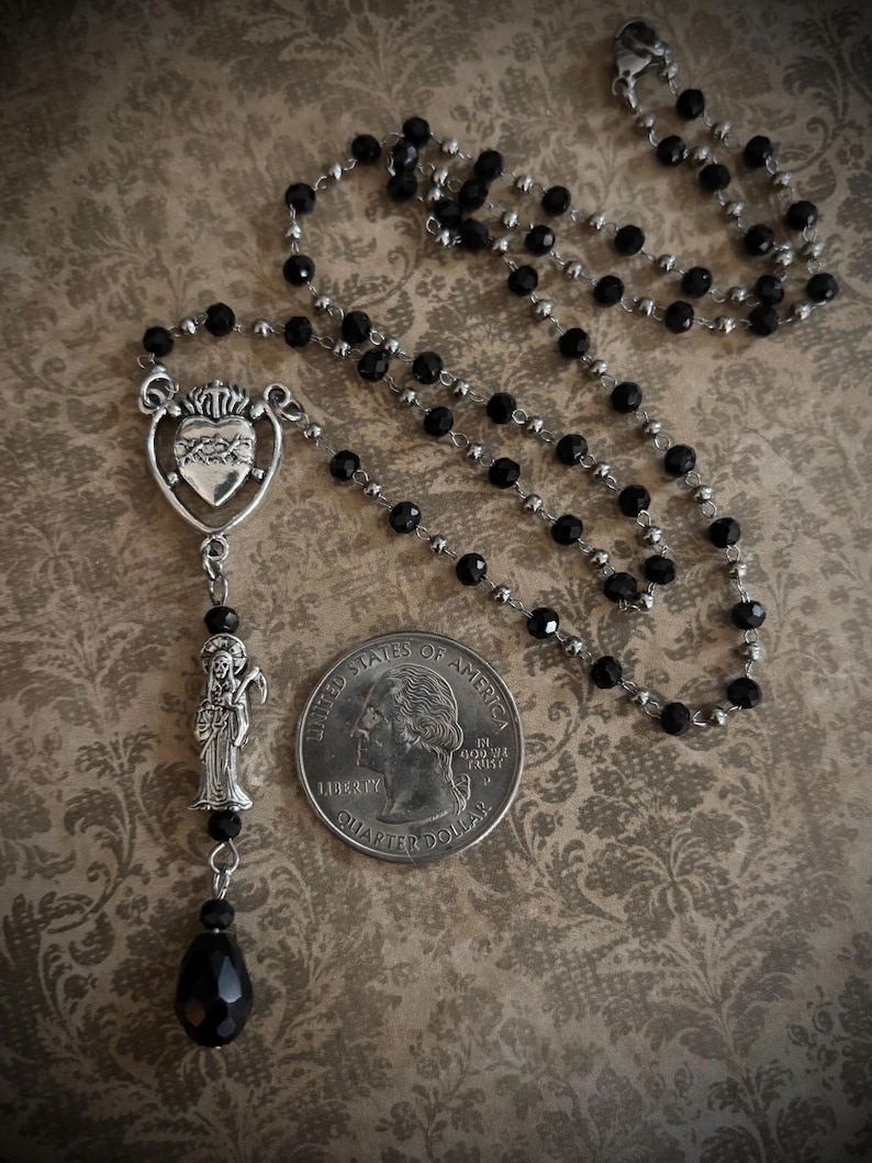Puede incluir: Un collar de rosario de cuentas negras con detalles met&aacute;licos plateados. El collar presenta un colgante de coraz&oacute;n con un dise&ntilde;o de llama, una figura de santo y una cuenta en forma de l&aacute;grima. Una moneda est&aacute; al lado del collar para comparar el tama&ntilde;o.