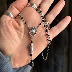 Puede incluir: Un collar de rosario plateado con cuentas negras. El colgante presenta un dise&ntilde;o de coraz&oacute;n con una corona y una peque&ntilde;a figura. Una gema negra en forma de l&aacute;grima cuelga en la parte inferior del colgante.