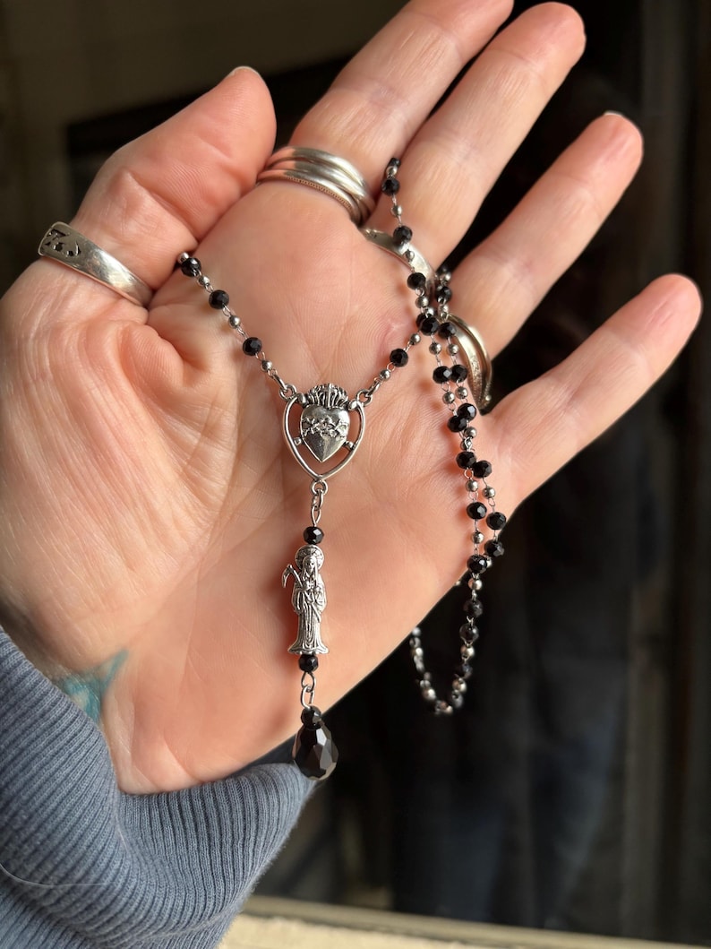Puede incluir: Un collar de rosario plateado con cuentas negras. El colgante presenta un dise&ntilde;o de coraz&oacute;n con una corona y una peque&ntilde;a figura. Una gema negra en forma de l&aacute;grima cuelga en la parte inferior del colgante.