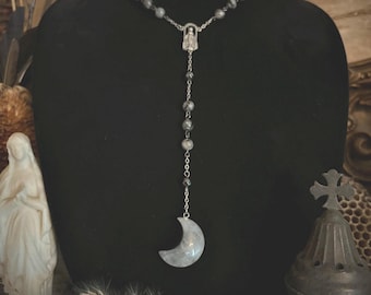 Puede incluir: Un collar de cadena de plata con un colgante de luna creciente y un pequeño amuleto religioso. El collar está hecho de cuentas grises y se muestra en un maniquí negro.