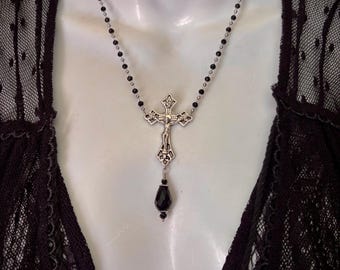 Puede incluir: Un collar estilo rosario con pequeñas cuentas negras intercaladas con detalles en metal plateado. La pieza central es un crucifijo plateado con una figura detallada, que cuelga sobre un colgante negro en forma de lágrima.