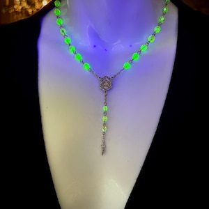 Puede incluir: Un collar de rosario de cuentas verdes brillantes con una pieza central y un colgante plateados. El collar se exhibe sobre un maniquí blanco, con un fondo negro. Las cuentas parecen luminosas.