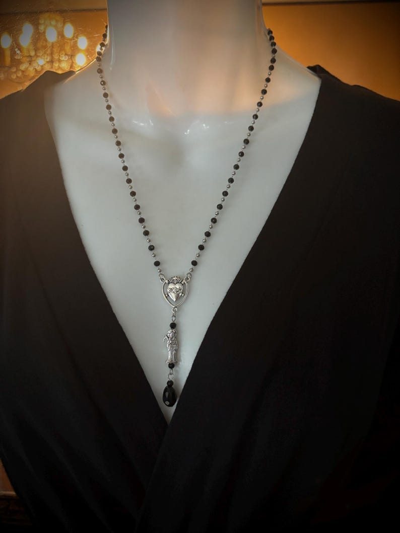 Puede incluir: Un collar largo con cuentas negras y detalles plateados. El colgante presenta un dise&ntilde;o en forma de coraz&oacute;n con una corona y una peque&ntilde;a figura. Una cuenta negra en forma de l&aacute;grima cuelga en la parte inferior del colgante. El collar se exhibe en un maniqu&iacute;.