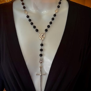 Puede incluir: Un collar de rosario con cuentas negras y detalles metálicos plateados. El collar presenta un colgante de crucifijo, dijes en forma de corazón y un medallón central con una figura religiosa. Las cuentas parecen ser de una piedra oscura.