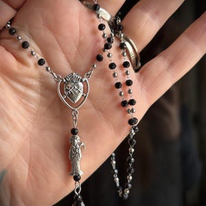 Puede incluir: Un collar plateado con cuentas negras y un colgante. El colgante presenta un coraz&oacute;n con llamas y una figura que sostiene una guada&ntilde;a. El collar se muestra sobre un fondo neutro.