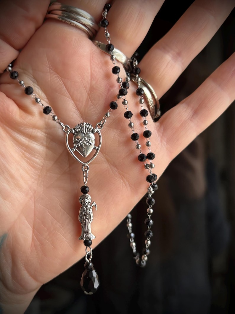 Puede incluir: Un collar plateado con cuentas negras y un colgante. El colgante presenta un coraz&oacute;n con llamas y una figura que sostiene una guada&ntilde;a. El collar se muestra sobre un fondo neutro.