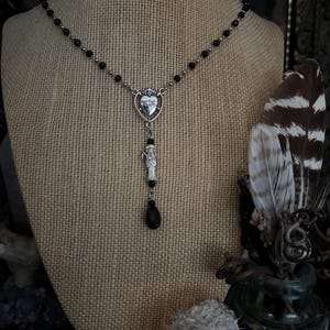 Puede incluir: Un collar plateado con un colgante en forma de coraz&oacute;n con una corona y una figura detallada. El collar tiene una cadena de cuentas con cuentas negras y un dije negro en forma de l&aacute;grima. El collar se exhibe en un maniqu&iacute;.