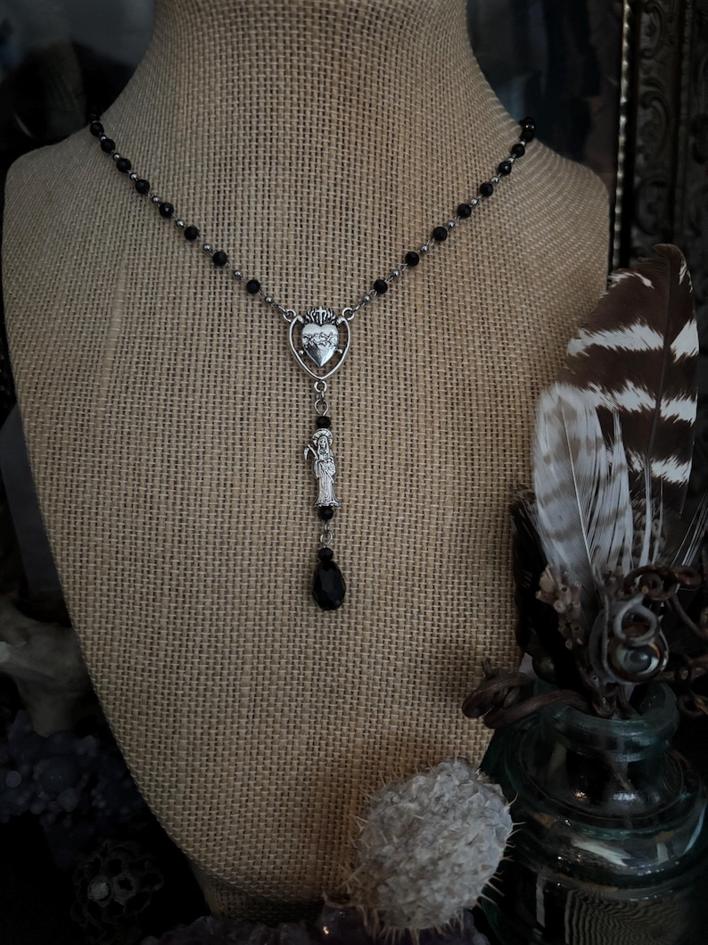 Puede incluir: Un collar plateado con un colgante en forma de coraz&oacute;n con una corona y una figura detallada. El collar tiene una cadena de cuentas con cuentas negras y un dije negro en forma de l&aacute;grima. El collar se exhibe en un maniqu&iacute;.
