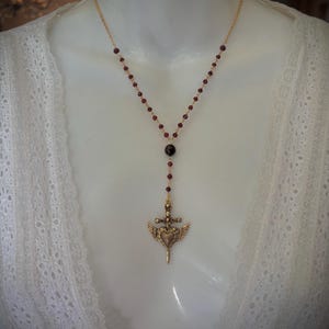 May include: A gold-toned necklace with a pendant featuring a heart, wings, and a sword. The necklace has a chain of small, dark red beads. The pendant is suspended from a longer strand of the same beads.