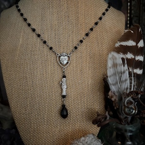 Puede incluir: Un collar plateado con cuentas negras y un colgante con un dise&ntilde;o de coraz&oacute;n y una figura de santo. El collar tiene un dije negro en forma de l&aacute;grima. El collar se exhibe en un maniqu&iacute; cubierto de arpillera.