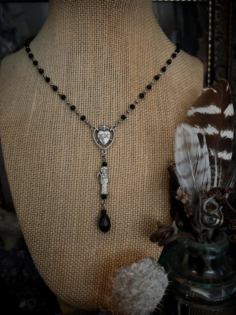 Puede incluir: Un collar plateado con cuentas negras y un colgante con un dise&ntilde;o de coraz&oacute;n y una figura de santo. El collar tiene un dije negro en forma de l&aacute;grima. El collar se exhibe en un maniqu&iacute; cubierto de arpillera.
