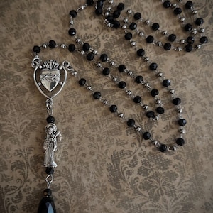 Puede incluir: Un rosario de cuentas negras con un colgante plateado. El colgante presenta un coraz&oacute;n con una corona y una figura de santo. El rosario tiene una cuenta en forma de l&aacute;grima en la parte inferior.