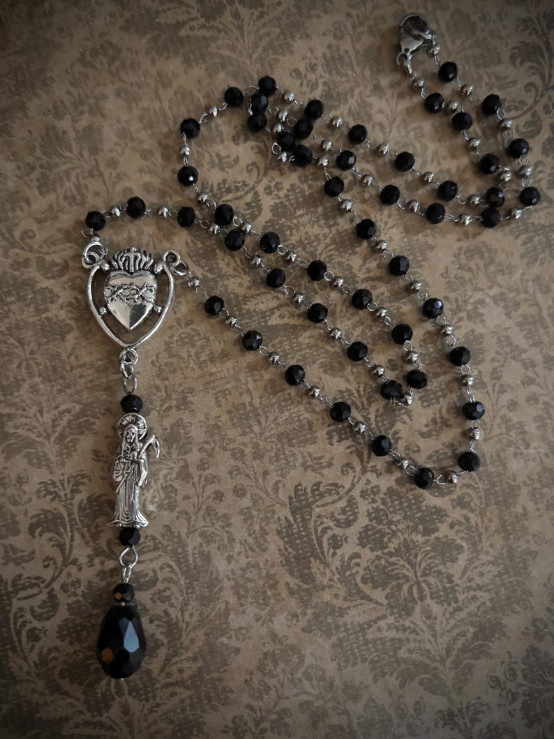 Puede incluir: Un rosario de cuentas negras con un colgante plateado. El colgante presenta un coraz&oacute;n con una corona y una figura de santo. El rosario tiene una cuenta en forma de l&aacute;grima en la parte inferior.
