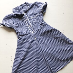 Puede incluir: Un vestido vintage a rayas azules y blancas con mangas cortas y cuello. El vestido presenta ribetes blancos a lo largo del cuello, bolsillo y tapeta de botones. El vestido tiene un corpi&ntilde;o ajustado y una falda acampanada.