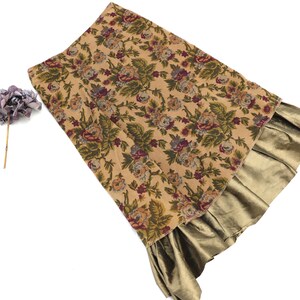 May include: A beige floral print skirt with a gold-coloured ruffle at the hem. The skirt features a pattern of roses and leaves in shades of green, purple, and brown. A purple flower is placed to the left of the skirt.