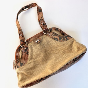May include: Tan handbag with brown snakeskin-patterned trim and handles. The bag features a central tan fabric panel, a zipper, and silver-toned hardware. The brand name "JAG" is visible on the front.