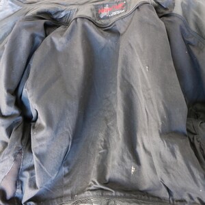 May include: Inside view of a black leather jacket with a black satin lining. The jacket is unzipped, revealing the interior construction and a small label at the neck. The lining shows some wear.