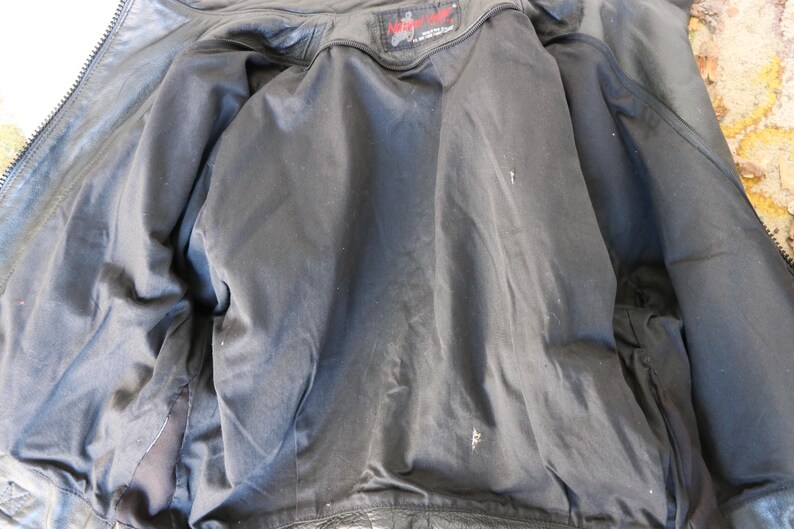 May include: Inside view of a black leather jacket with a black satin lining. The jacket is unzipped, revealing the interior construction and a small label at the neck. The lining shows some wear.