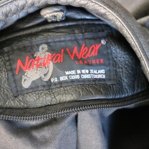 May include: Close-up of a black leather jacket with a visible label. The label reads "Natural Wear Leather" in red, with a motorcycle graphic. The text "MADE IN NEW ZEALAND" and address details are also visible.