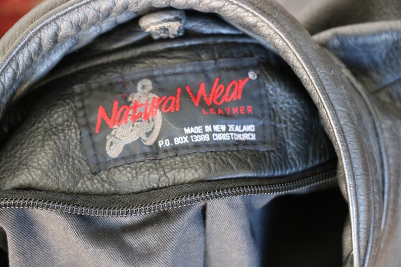 May include: Close-up of a black leather jacket with a visible label. The label reads "Natural Wear Leather" in red, with a motorcycle graphic. The text "MADE IN NEW ZEALAND" and address details are also visible.