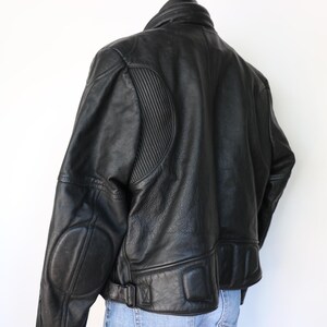 May include: Black leather motorcycle jacket with a ribbed design on the shoulders and padded elbow patches. The jacket has a collar, a belt at the waist, and a zipper closure. The jacket is worn over blue jeans.