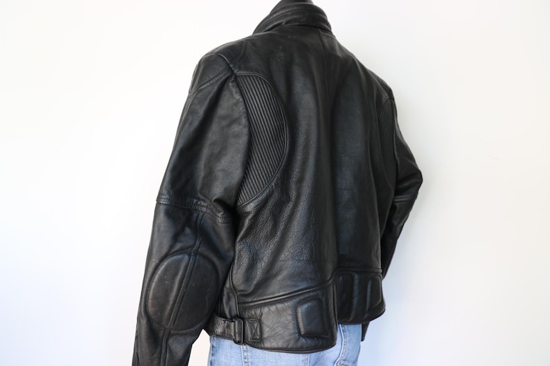 May include: Black leather motorcycle jacket with a ribbed design on the shoulders and padded elbow patches. The jacket has a collar, a belt at the waist, and a zipper closure. The jacket is worn over blue jeans.