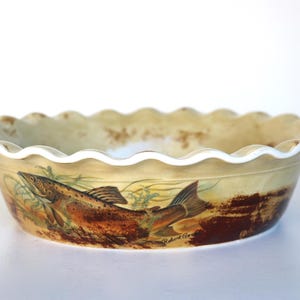 May include: A round ceramic pie dish with a scalloped rim, featuring a beige and brown colour palette. The exterior displays a detailed illustration of a brown trout swimming among aquatic plants, with the signature Robert Gordon printed near the tail.