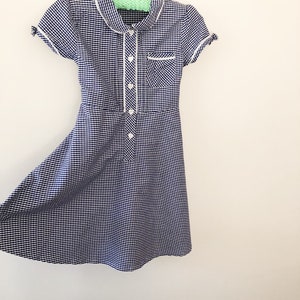 Puede incluir: Un vestido de cuadros vichy azul marino y blanco con cuello blanco, ribete de bolsillo y detalles de botones. El vestido de manga corta est&aacute; colgado de una percha de madera sobre un fondo blanco. El vestido tiene un corpi&ntilde;o entallado y una falda acampanada.