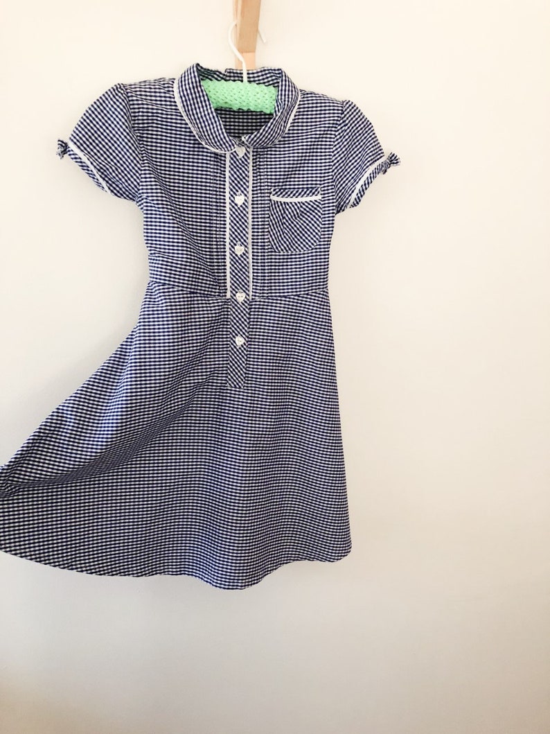 Puede incluir: Un vestido de cuadros vichy azul marino y blanco con cuello blanco, ribete de bolsillo y detalles de botones. El vestido de manga corta est&aacute; colgado de una percha de madera sobre un fondo blanco. El vestido tiene un corpi&ntilde;o entallado y una falda acampanada.