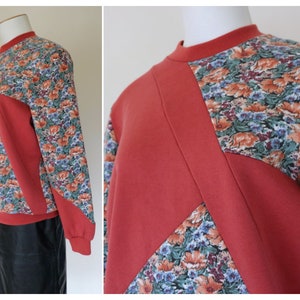 May include: A red sweatshirt with a geometric pattern of floral fabric. The sleeves and a portion of the front and back are made of a floral fabric with orange, blue, and green flowers.