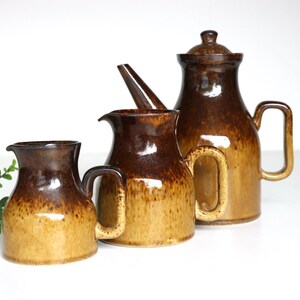 May include: A collection of three ceramic jugs in brown and tan hues. The largest jug has a lid. The jugs feature handles and a glossy finish. The set is displayed on a white surface.