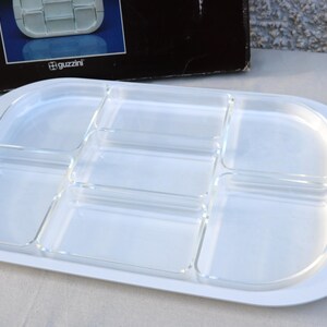 May include: White serving tray with six clear compartments. The tray has a rounded rectangular shape with a white border and a textured design on the sides. The Guzzini logo is visible on the box.