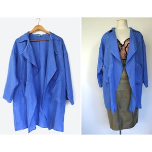 May include: A long, open-front blue coat with a wide lapel and pockets. The coat is displayed on a wooden hanger and also on a mannequin. The coat is a solid blue colour and appears to be made of a lightweight fabric.