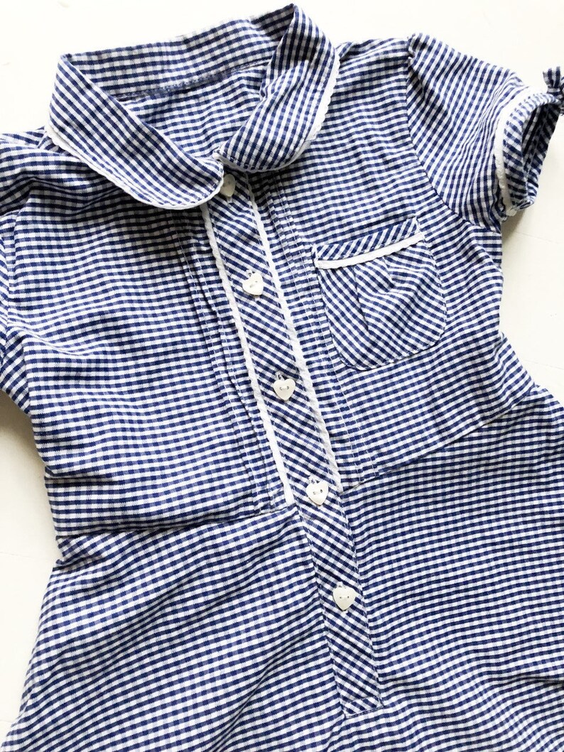 Puede incluir: Vestido a cuadros azul y blanco con cuello y ribete blancos. El vestido presenta botones en forma de coraz&oacute;n y un peque&ntilde;o bolsillo. Las mangas cortas est&aacute;n rematadas con un ribete blanco y peque&ntilde;os lazos. El vestido es de tejido ligero.