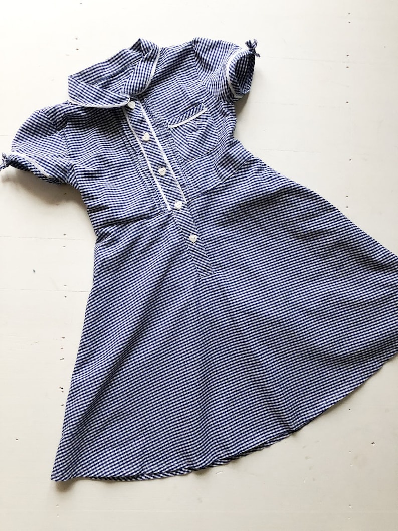 Puede incluir: Un vestido vintage a rayas azules y blancas con mangas cortas y cuello. El vestido presenta ribetes blancos a lo largo del cuello, bolsillo y tapeta de botones. El vestido tiene un corpi&ntilde;o ajustado y una falda acampanada.