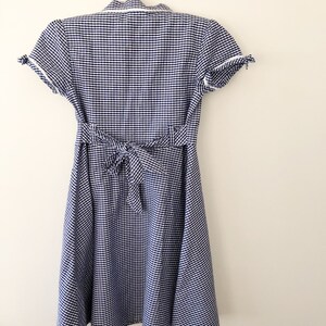 Puede incluir: Un vestido de cuadros vichy azul y blanco con mangas cortas y cintura anudada en la espalda. El vestido est&aacute; colgado de una percha blanca contra una pared blanca. El vestido tiene cuello y ribete blanco en las mangas.