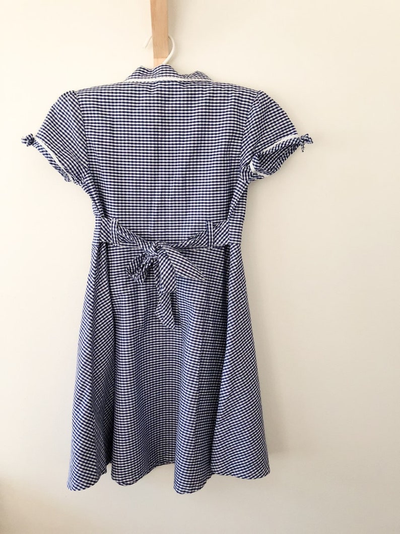 Puede incluir: Un vestido de cuadros vichy azul y blanco con mangas cortas y cintura anudada en la espalda. El vestido est&aacute; colgado de una percha blanca contra una pared blanca. El vestido tiene cuello y ribete blanco en las mangas.