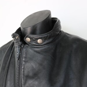 May include: Close-up of a black leather jacket with a stand-up collar and a zipper closure. The jacket has silver-colored snap details on the collar and a smooth, textured appearance. The jacket is displayed on a black mannequin.