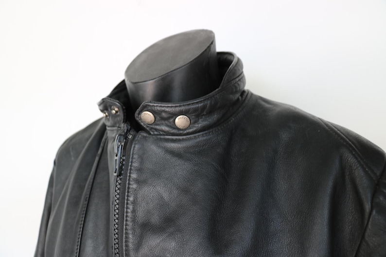 May include: Close-up of a black leather jacket with a stand-up collar and a zipper closure. The jacket has silver-colored snap details on the collar and a smooth, textured appearance. The jacket is displayed on a black mannequin.