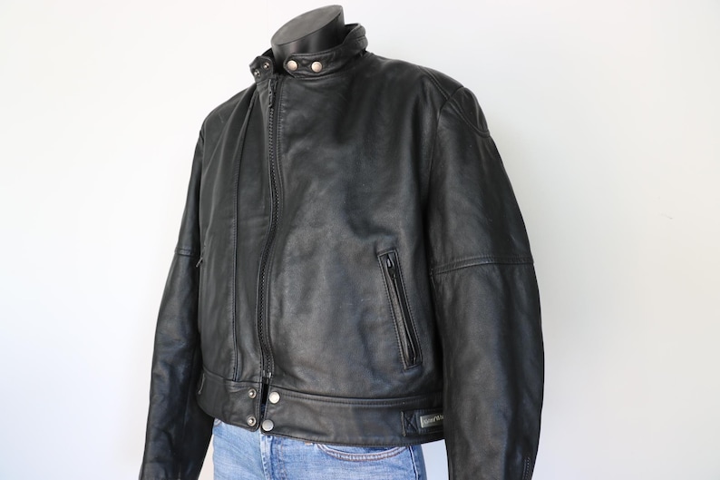 May include: A black leather jacket with a zip-up front, a small pocket on the chest, and a round collar. The jacket has a classic design and is displayed on a mannequin.