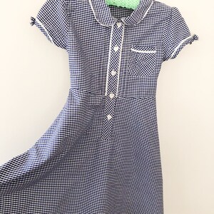Puede incluir: Un vestido de cuadros vichy azul marino y blanco con cuello Peter Pan y mangas cortas. El vestido presenta ribetes blancos a lo largo del cuello, el bolsillo y la tapeta de botones. Tiene un corpi&ntilde;o entallado y una falda acampanada, colgado de una percha de madera.