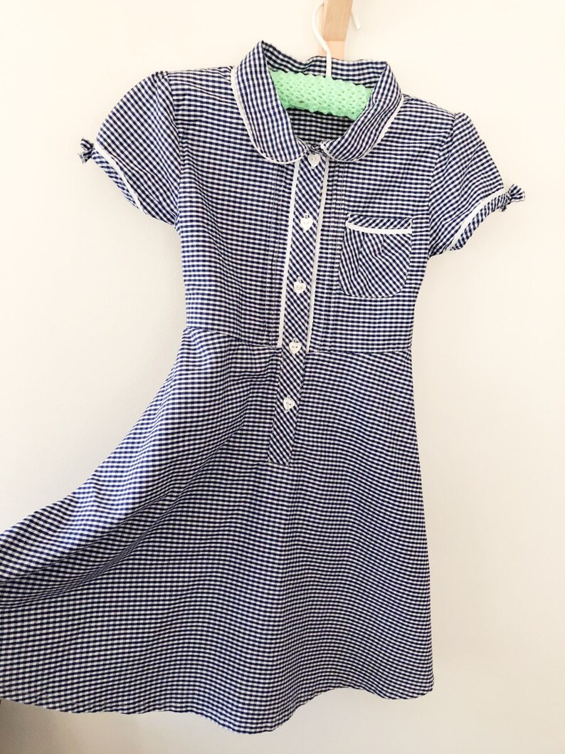 Puede incluir: Un vestido de cuadros vichy azul marino y blanco con cuello Peter Pan y mangas cortas. El vestido presenta ribetes blancos a lo largo del cuello, el bolsillo y la tapeta de botones. Tiene un corpi&ntilde;o entallado y una falda acampanada, colgado de una percha de madera.