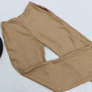 May include: A pair of tan trousers with a loose fit, placed on a white surface. A black fedora hat with a dark band is next to the trousers. The trousers seem to be made of a light material.