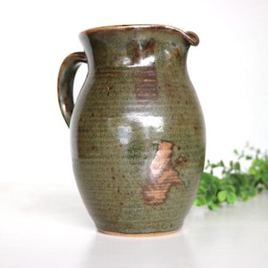 May include: A handcrafted, green ceramic jug with a handle and spout. The jug has a rustic, textured appearance with brown accents. It is a decorative item for the kitchen or dining room.