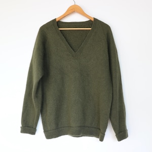 May include: An olive green V-neck sweater with long sleeves. The sweater has a soft, textured appearance and features ribbed cuffs and hem. It is displayed on a wooden hanger against a white backdrop.