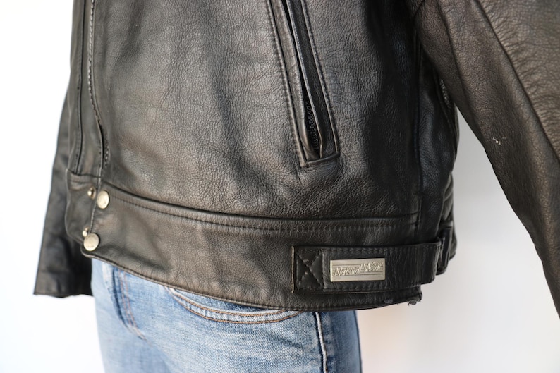 May include: A black leather jacket with a zipper pocket and silver buttons. The jacket has a belt detail at the waist and a small rectangular metal tag that reads "Roper Rider".