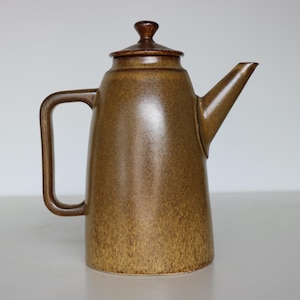 May include: A brown ceramic coffee pot with a lid and handle. The pot has a spout for pouring and a rounded body. The color is a speckled brown with a darker brown handle and lid. The pot is likely vintage or retro.