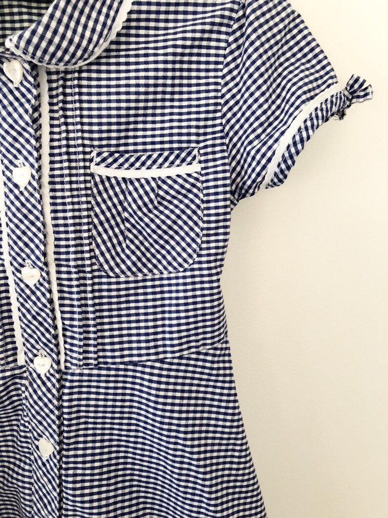 Puede incluir: Vestido de cuadros azules y blancos con cuello, ribete de bolsillo y pu&ntilde;os blancos. El vestido tiene una parte delantera abotonada con botones en forma de coraz&oacute;n. Las mangas cortas est&aacute;n rematadas con ribete blanco.